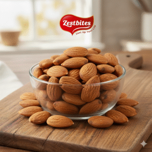 A clear glass bowl filled with Zestbites Jumbo California Almonds sits on a rustic wooden cutting board, with some almonds scattered around the base of the bowl. The Zestbites logo is visible at the top center of the image, against a subtly blurred background of a kitchen setting.